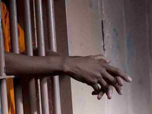 Cropped view of a prisoner in an orange jumpsuit standing in a prison cell with arms extended through the bars. Horizontal format.