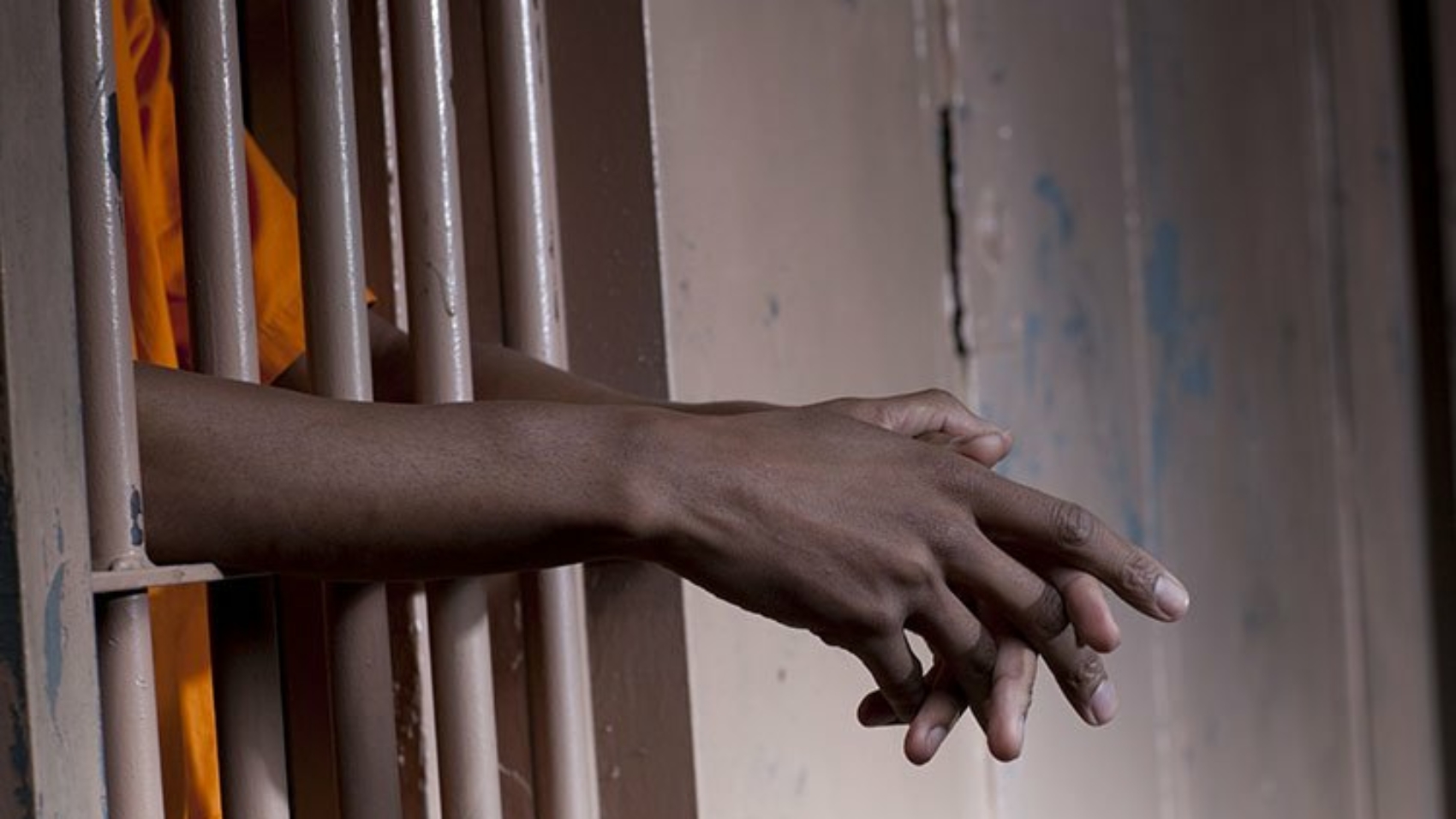 Cropped view of a prisoner in an orange jumpsuit standing in a prison cell with arms extended through the bars. Horizontal format.