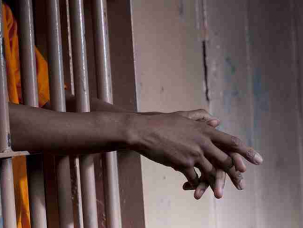 Cropped View Of A Prisoner In An Orange Jumpsuit Standing In A Prison Cell With Arms Extended Through The Bars. Horizontal Format.